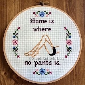 Peut inclure: Un cerceau de broderie en point de croix blanc avec un cadre en bois brun. La broderie représente une paire de jambes dans une pose détendue avec le texte "Home is where no pants is."