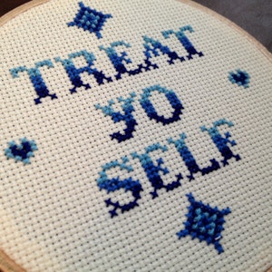 May include: Cross-stitch embroidery hoop with the phrase "TREAT YO SELF" in varying shades of blue. The design includes decorative blue starburst and heart shapes on a white fabric background.