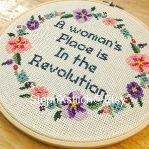 Peut inclure: Un cercle de broderie blanc avec un motif de point de croix d'une couronne de fleurs entourant le texte "A woman's place is in the revolution".