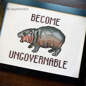 May include: Cross-stitch art in a black frame. The artwork features a pixelated hippo with the words "BECOME UNGOVERNABLE" in pink. The hippo is gray, brown, and pink, with visible cross-stitch texture.