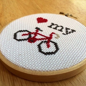 Puede incluir: Un aro de bordado blanco con un corazón rojo, una bicicleta roja y el texto "Me encanta mi" bordado en él.