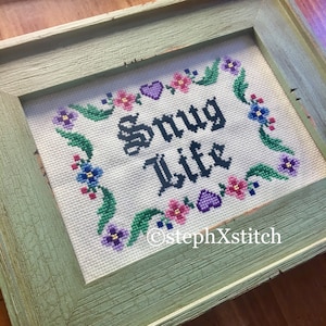 Könnte beinhalten: Ein gerahmtes Kreuzstichbild mit dem Text "Snug Life" in schwarzen Buchstaben, umgeben von einem Rand aus grünen Blättern und bunten Blumen. Der Rahmen ist aus hellgrünem Holz mit einer abgenutzten Oberfläche.