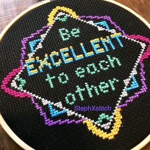 Puede incluir: Un aro de bordado negro con un diseño de punto de cruz colorido. El diseño presenta una forma de estrella con el texto "Be excellent to each other" en un arcoíris de colores. El nombre del artista, "StephXStitch", está bordado en la esquina inferior.