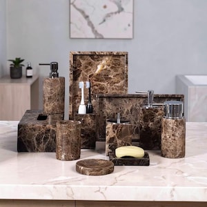 Op de afbeelding: Een verzameling bruinmarmeren badkameraccessoires, waaronder een zeepdispenser, een tissuebox, een tandenborstelhouder, een dienblad, een beker, een zeepbakje en een lotiondispenser. De set is geplaatst op een lichtkleurig aanrecht.