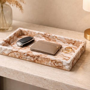 Può includere: Un vassoio decorativo rettangolare con un motivo marmorizzato contiene una chiave dell'auto, un portafoglio e due anelli d'oro. Il vassoio è su una superficie chiara, con una lampada e un vaso di fiori secchi sullo sfondo.