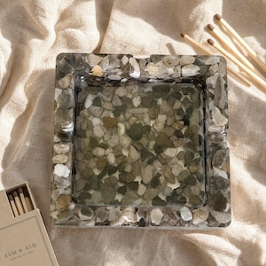 May include: A square ashtray made of clear resin embedded with various small, smooth, multi-colored river stones in shades of gray, tan, and white. The ashtray features four notches on the edges and a flat, transparent base filled with stones.