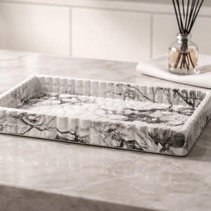 Può includere: Vassoio rettangolare effetto marmo con un motivo bianco e grigio. Il vassoio ha un bordo strutturato e scanalato. Sullo sfondo un flacone di vetro con canne e un asciugamano bianco piegato.