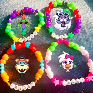May include: Four colorful beaded bracelets with character charms. One bracelet says "Monty" and features a green alligator charm. Another says "Roxy" with a wolf charm. The third says "Freddy" with a bear charm, and the last says "Chica" with a chicken charm.