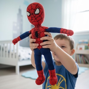 以下が含まれることがあります： 赤、青、黒のハンドメイドかぎ針編みスパイダーマン人形。人形は赤い部分に詳細なウェブパターンと白い目のディテールがあります。人形は、黄色のクモのグラフィックが入った青いTシャツを着た人が持っています。