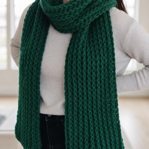 Puede incluir: Una bufanda gruesa de punto verde bosque. La bufanda est&aacute; envuelta alrededor del cuello y cuelga por delante. El patr&oacute;n de punto es visible y la bufanda parece suave y c&aacute;lida. La persona lleva un su&eacute;ter gris claro y vaqueros oscuros.