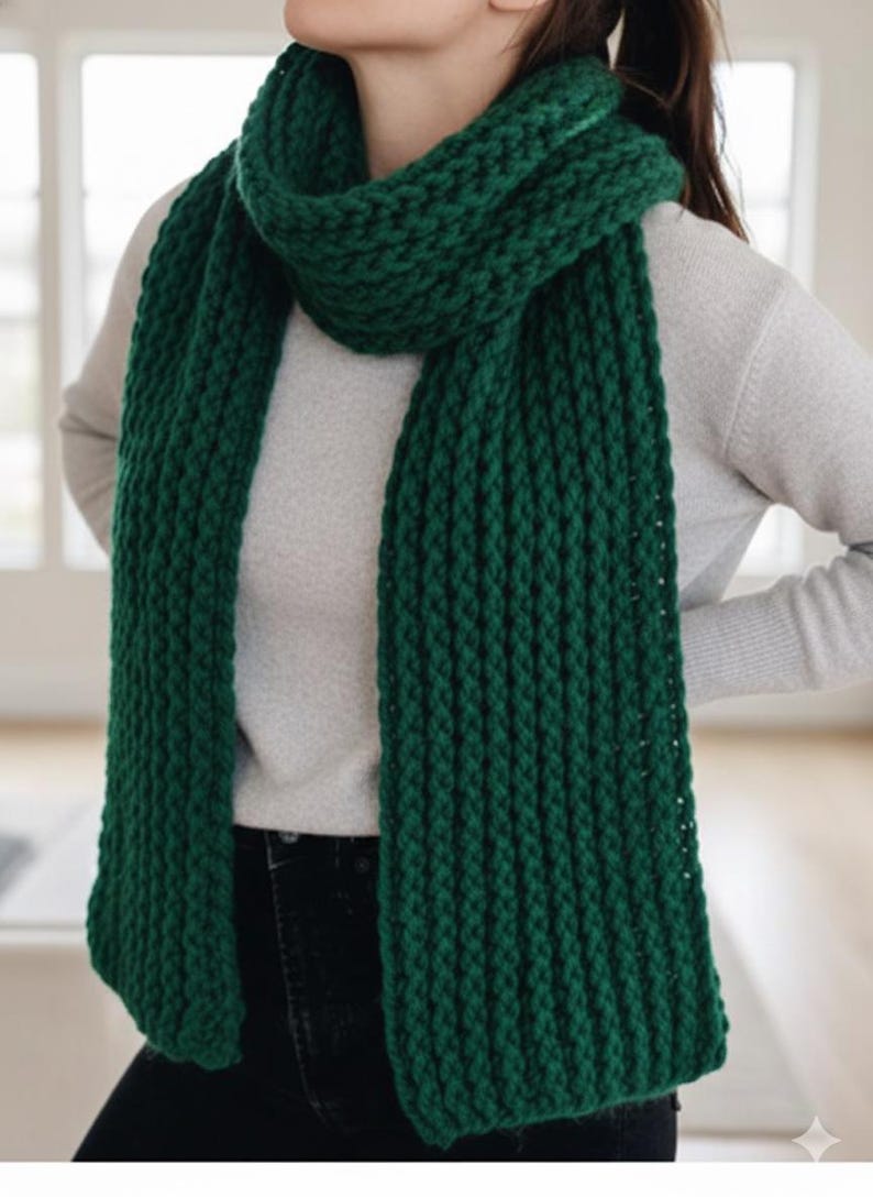 Puede incluir: Una bufanda gruesa de punto verde bosque. La bufanda est&aacute; envuelta alrededor del cuello y cuelga por delante. El patr&oacute;n de punto es visible y la bufanda parece suave y c&aacute;lida. La persona lleva un su&eacute;ter gris claro y vaqueros oscuros.