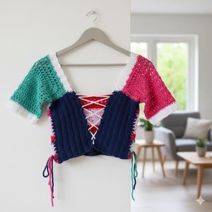 Puede incluir: Top de ganchillo colorido con mangas cortas, colgado de una percha gris. El top presenta un dise&ntilde;o de patchwork con secciones turquesa, rosa, azul marino y blanco. La espalda tiene un detalle de cordones con cordones rosas y blancos.