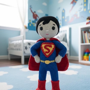 Op de afbeelding: Gehaakte Superman-pop in blauw pak met rode cape en laarzen. De pop heeft een gele riem en een borstembleem met een rood hart en een gele "S". De pop staat op een blauw tapijt met een wolkenpatroon.
