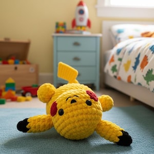 Könnte beinhalten: Handgefertigtes gelbes Pikachu-Plüschtier aus Häkelarbeit mit schwarzen Augen, roten Wangen und schwarzspitzigen Armen und Füßen. Das Kuscheltier liegt auf einem blauen Teppich in einem Kinderzimmer. Ein gelber Schwanz und Ohren sind sichtbar.