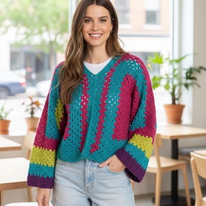 Puede incluir: Un su&eacute;ter de crochet colorido con cuello en V. El su&eacute;ter presenta un patr&oacute;n vibrante de turquesa, fucsia, amarillo y morado. Las mangas son largas y acampanadas, y el su&eacute;ter se combina con pantalones cortos de mezclilla. El estilo general es casual y hecho a mano.