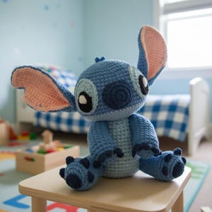 Könnte beinhalten: Handgemachte blaue Stitch-Häkelpuppe mit großen Ohren, schwarzen Augen und einem hellblauen Körper. Die Puppe sitzt auf einem kleinen Holztisch in einem Kinderzimmer. Die Ohren sind innen rosa.