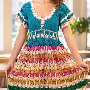 Puede incluir: Vestido de ganchillo colorido con escote en V. La parte superior es turquesa con ribete crema, que se transforma en una falda con rayas horizontales rosas, beige y doradas. El dobladillo est&aacute; terminado con ganchillo verde.