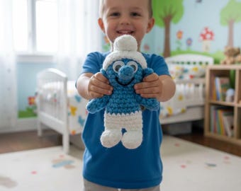 Op de afbeelding: Een handgemaakte gehaakte Smurf-pop, met een blauw lichaam, witte broek en een witte muts. De pop heeft grote, ronde ogen en een glimlachende uitdrukking. De pop wordt vastgehouden door een kind.