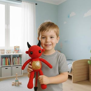 May include: A red crocheted devil doll with black horns and a gold sequined belly. The handmade plush toy has a long tail and is being held by a child. The doll is a unique, whimsical item.