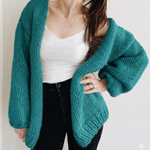 Puede incluir: Un c&aacute;rdigan de punto grueso de color verde azulado con mangas largas. El su&eacute;ter abierto est&aacute; hecho de un hilo grueso y tiene un ajuste relajado. La modelo lleva una camiseta sin mangas blanca y vaqueros negros.