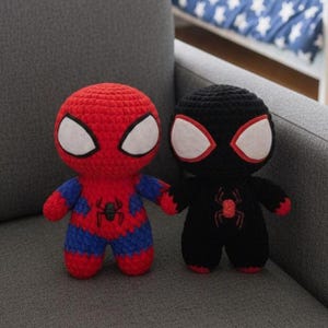 Puede incluir: Dos muñecos de superhéroes tejidos a ganchillo. Uno es rojo con detalles azules y un emblema de araña, el otro es negro con un emblema de araña rojo. Ambos muñecos tienen forma de ojos blancos y están sobre una superficie gris.
