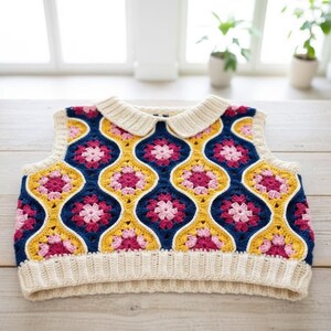 Puede incluir: Un chaleco de ganchillo hecho a mano y colorido, con cuello y ribetes color crema. El chaleco presenta un patr&oacute;n repetitivo de flores rosas y amarillas sobre un fondo azul marino. El chaleco se exhibe sobre una superficie de madera.