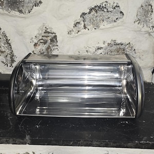 以下が含まれることがあります： カーブしたロールトップの蓋が付いた、光沢のあるステンレス製のパンボックス。ボックスは長方形で、丸みを帯びたエッジと反射面があります。内部が見え、ボックスは空です。パンボックスは黒い表面に置かれています。