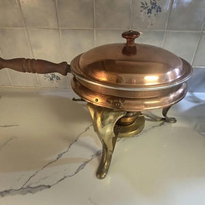 May include: A copper chafing dish with a lid and wooden handle. The dish sits on a brass stand with three decorative legs. The lid has a small, round wooden knob.
