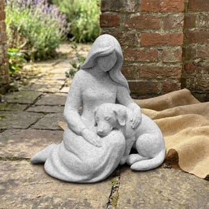 May include: A light gray speckled stone-effect sculpture depicting a person with long hair sitting on the ground while gently embracing a dog. The figure is wearing a long garment that covers their legs, and the dog is resting beside them.
