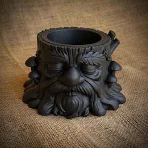 May include: Black planter shaped like a tree stump with a carved face, leaves, and mushrooms. The planter has a wide opening and a textured surface. The design features a serene face with closed eyes and a long beard.
