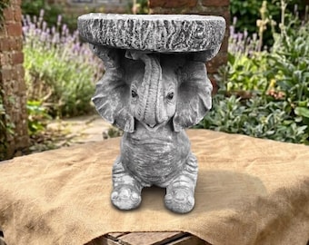 May include: A gray stone-colored garden stool shaped like a seated elephant. The elephant has large ears, a trunk curled upward, and textured skin. It supports a flat, circular, rough-textured top surface that serves as a small table.