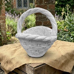 May include: A light gray, stone-like basket featuring a woven texture on the main body and a thick, braided handle. The basket has a rounded, open top and a sturdy base, designed with a consistent matte finish that mimics the appearance of concrete.