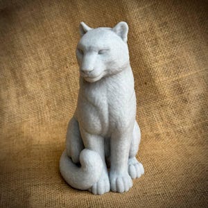 May include: A light gray sculpture of a seated feline with its tail curled. The statue has detailed fur texture and is posed with its head slightly lowered. The sculpture is set against a neutral background.