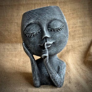 May include: A gray decorative planter shaped like a face with closed eyes and a finger pressed to the lips. The sculpture has a textured finish and is designed to hold small plants or flowers. The overall theme is a serene, artistic piece.