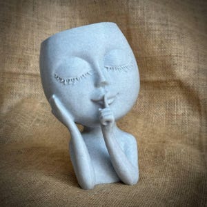 May include: A light blue decorative planter shaped like a face with closed eyes and a finger pressed to the lips. The planter has a textured surface and is designed to hold small plants or flowers. The overall theme is a serene, artistic piece.