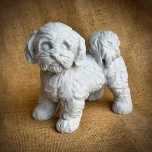 May include: A light gray, stone-like sculpture of a small dog, possibly a Shih Tzu or similar breed. The dog is standing with its head slightly turned, showcasing detailed fur texture and facial features. The sculpture is on a brown, textured background.