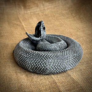 May include: A coiled gray snake sculpture with its mouth open, revealing a dark interior. The snake's body is textured with a scale pattern. The sculpture is set against a neutral, textured background.