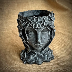 May include: A gray planter shaped like a woman's face, adorned with a floral crown and flowing hair. The detailed sculpture has a textured finish, creating a vintage aesthetic. Ideal for small plants or as a decorative piece.