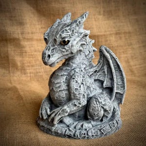 May include: A detailed, gray dragon figurine with textured scales, wings, and a contemplative expression. The dragon is seated, with its forepaws resting on its base. The base has a decorative border. The dragon's eyes are a warm, golden color.