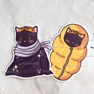 May include: Two stickers featuring cartoon cats. One is a black cat wearing goggles and a scarf, with stars. The other is a black cat in a yellow sleeping bag. The stickers are on a textured, off-white background.
