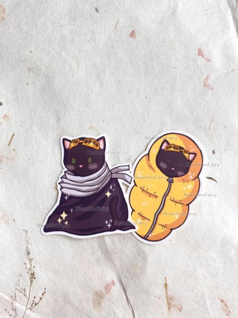 May include: Two stickers featuring cartoon cats. One is a black cat wearing goggles and a scarf, with stars. The other is a black cat in a yellow sleeping bag. The stickers are on a textured, off-white background.