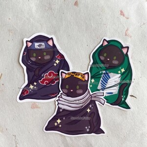 May include: Three cat stickers with anime-inspired designs. One cat wears a ninja headband and a dark purple cloak with red cloud accents. Another cat is in a green cloak with a white emblem. The third cat wears goggles and a scarf.
