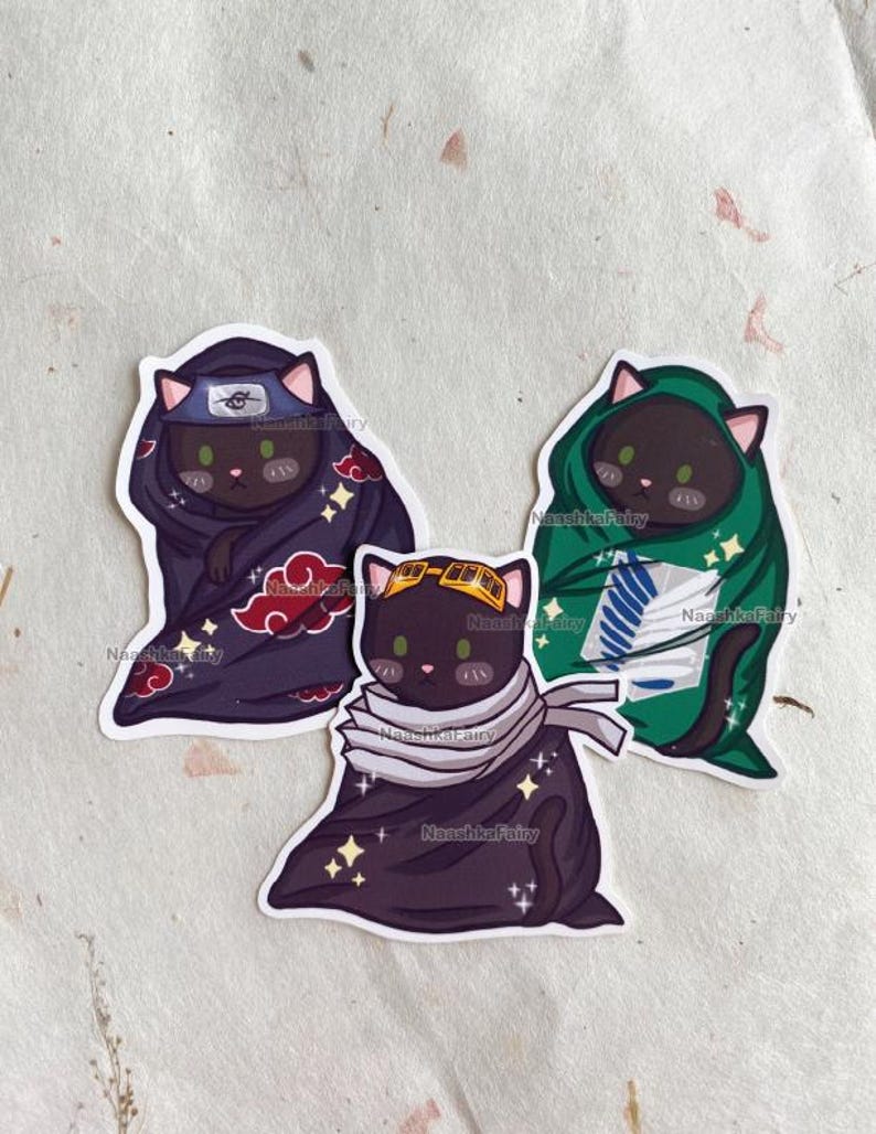 May include: Three cat stickers with anime-inspired designs. One cat wears a ninja headband and a dark purple cloak with red cloud accents. Another cat is in a green cloak with a white emblem. The third cat wears goggles and a scarf.