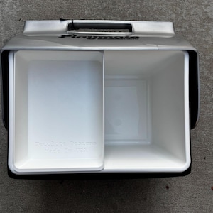 May include: A white and silver Playmate cooler with a black handle and trim. The cooler has two compartments, one with a sliding tray. The text "Recolote Designs Made In USA" is visible on the tray.