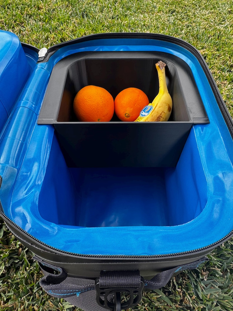 Puede incluir: Bolsa nevera negra y azul abierta con un compartimento interior negro. Dos naranjas y un pl&aacute;tano est&aacute;n dentro del compartimento. La nevera est&aacute; sobre hierba verde.