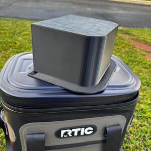 Puede incluir: Un accesorio rectangular gris para nevera se encuentra encima de una nevera RTIC negra y gris. El accesorio tiene una parte superior redondeada y parece estar impreso en 3D. La nevera est&aacute; sobre hierba, con una calle y casas al fondo.