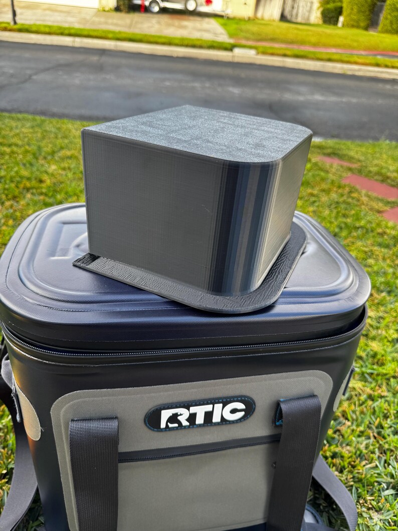 Puede incluir: Un accesorio rectangular gris para nevera se encuentra encima de una nevera RTIC negra y gris. El accesorio tiene una parte superior redondeada y parece estar impreso en 3D. La nevera est&aacute; sobre hierba, con una calle y casas al fondo.