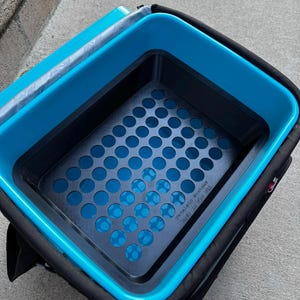May include: A rectangular storage container with a bright blue interior and a black exterior. The base features a grid of circular cutouts, and the text "Geneless Designs" is embossed in the bottom corner of the black plastic insert.