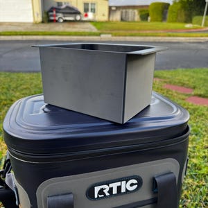 Puede incluir: Una nevera port&aacute;til RTIC negra y gris con un contenedor rectangular gris a juego encima. La nevera tiene una tapa negra y un panel frontal gris con el logotipo de RTIC. La escena es al aire libre con un fondo residencial.