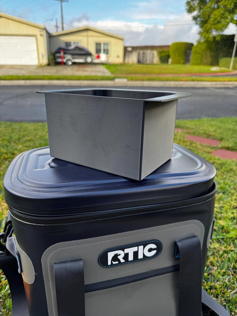 Puede incluir: Una nevera port&aacute;til RTIC negra y gris con un contenedor rectangular gris a juego encima. La nevera tiene una tapa negra y un panel frontal gris con el logotipo de RTIC. La escena es al aire libre con un fondo residencial.